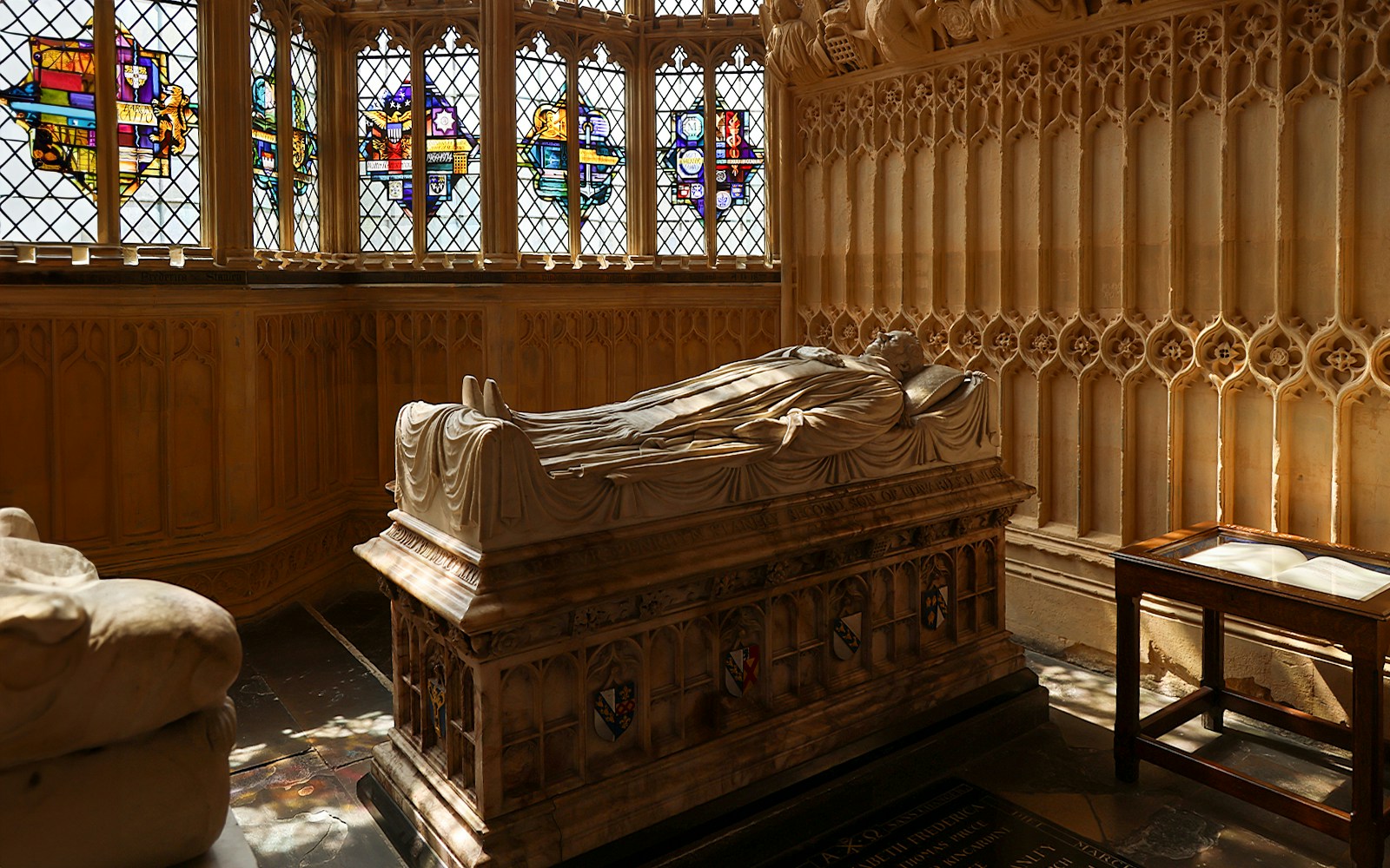 Tomb with effigy inside Westminster Abbey, London, with stained glass windows in the background.