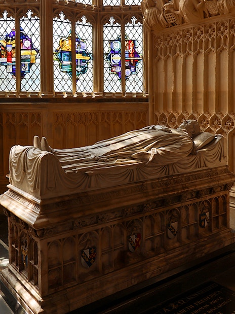 Tomb with effigy inside Westminster Abbey, London, with stained glass windows in the background.