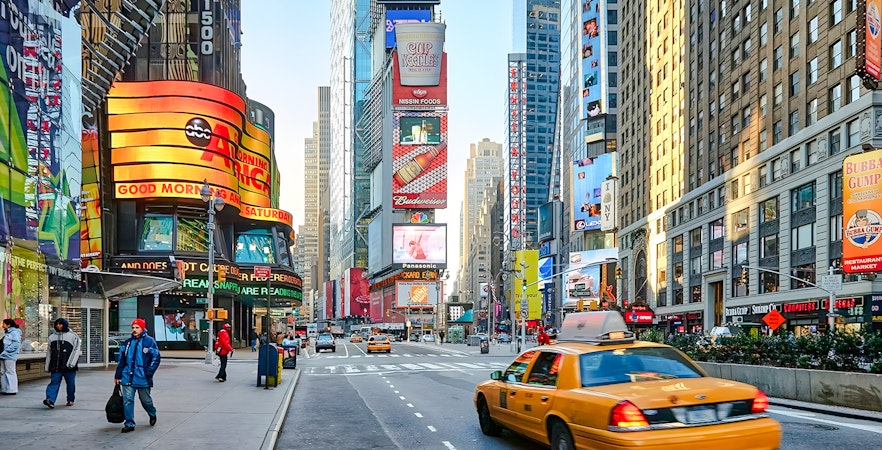 Yellow taxi driving through Times Square, New York City, surrounded by bright billboards and skyscrapers.