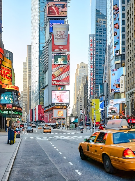 Yellow taxi driving through Times Square, New York City, surrounded by bright billboards and skyscrapers.
