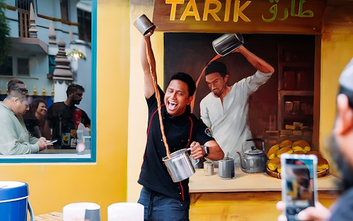 Person skillfully pouring Teh Tarik at a vibrant stall, with onlookers watching.