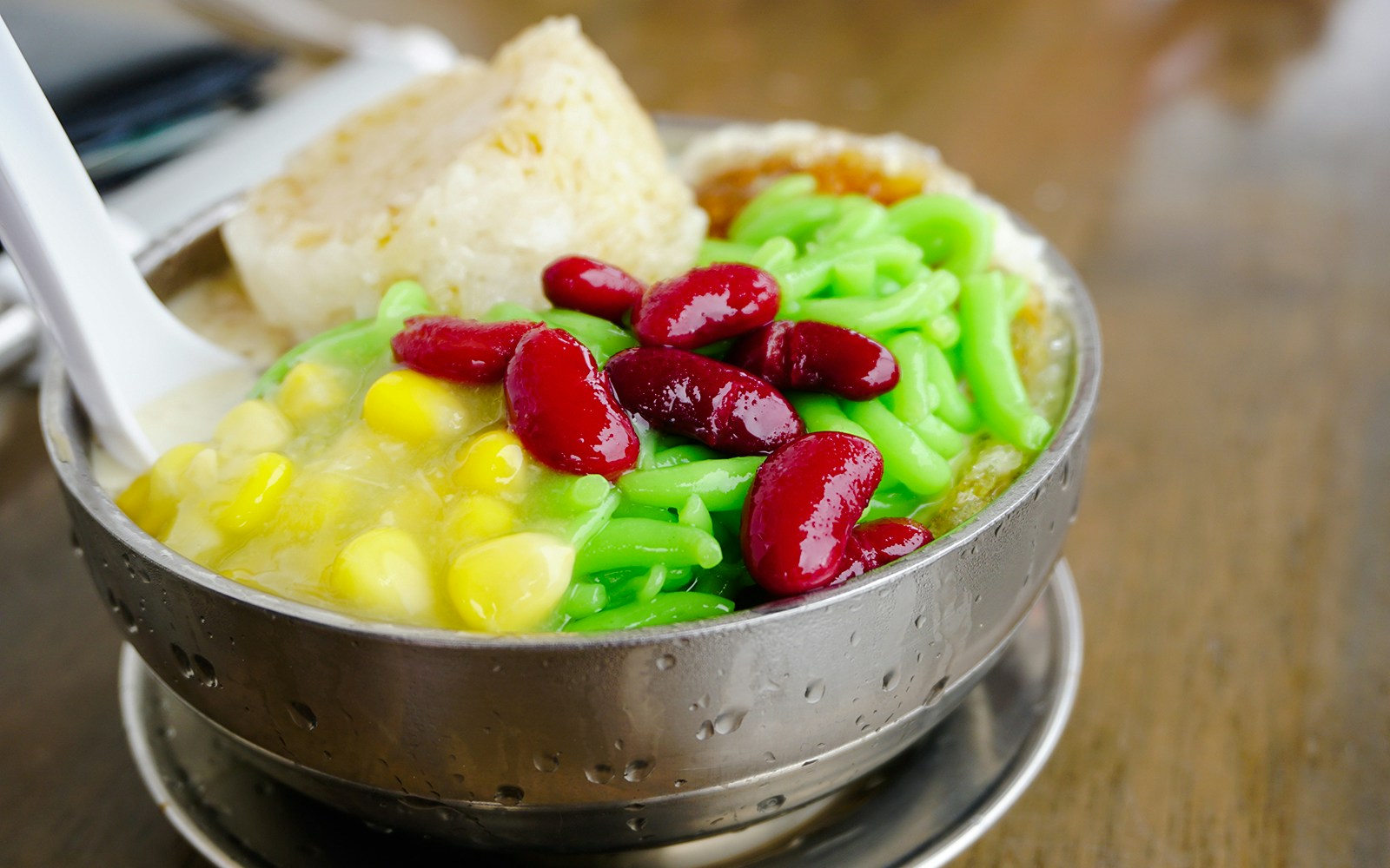 Chendol dessert with green jelly noodles, red beans, and corn in a metal bowl.