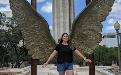 Person posing with large wing sculpture in San Antonio during city exploration game.