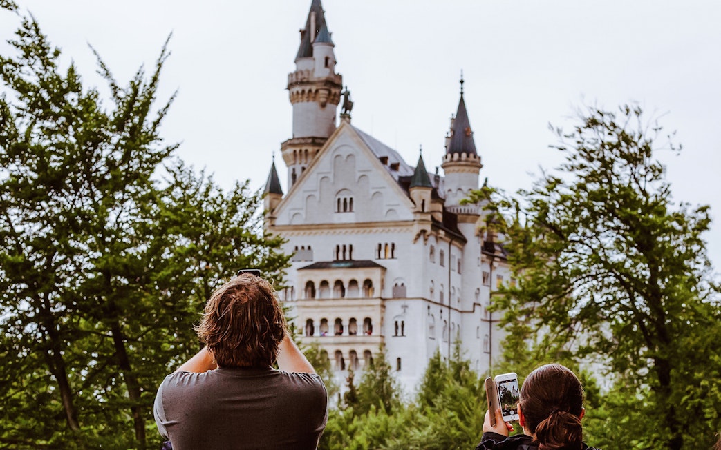 Visitors photographing Neuschwanstein Castle through trees in Bavaria, Germany.