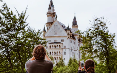 Visitors photographing Neuschwanstein Castle through trees in Bavaria, Germany.