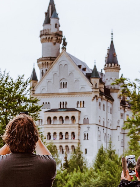 Visitors photographing Neuschwanstein Castle through trees in Bavaria, Germany.