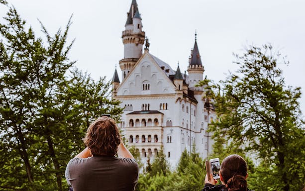 Visitors photographing Neuschwanstein Castle through trees in Bavaria, Germany.