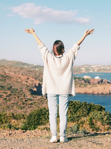Woman with arms raised on Cape Sounion cliff overlooking the Aegean Sea, Greece.