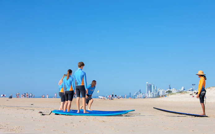 Surf instructor teaching beginners on Gold Coast beach with city skyline in background.