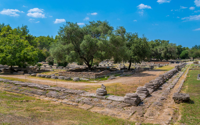 Leonidaion ruins at Olympia archaeological site, Greece, surrounded by trees and ancient stone structures.