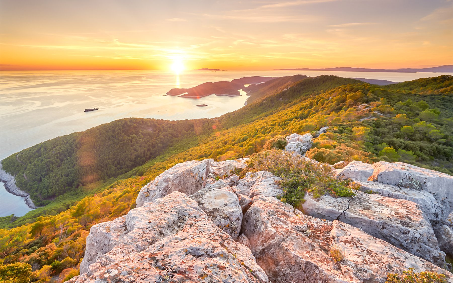 Dubrovnik coastline at sunset with rocky foreground and lush green hills.