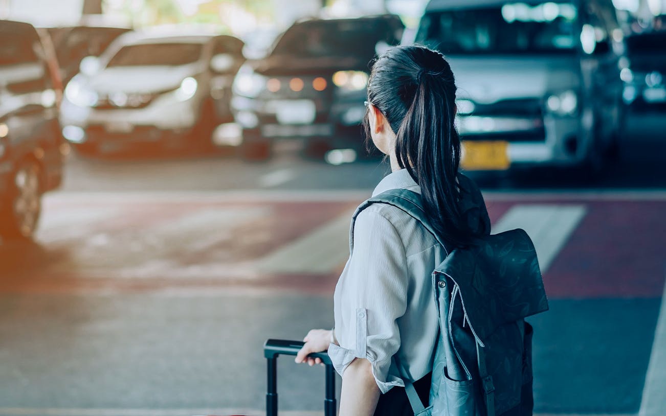 Woman with suitcase waiting for airport transfer, taxis in background.
