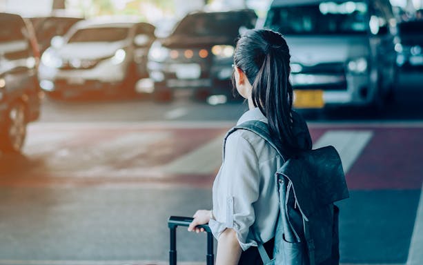 Woman with suitcase waiting for airport transfer, taxis in background.