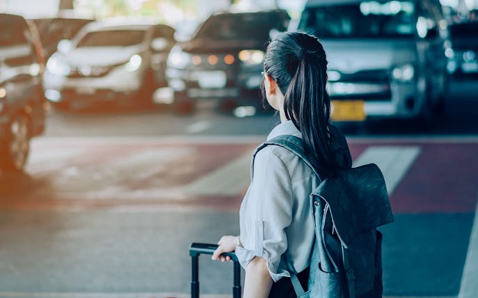 Woman with suitcase waiting for airport transfer, taxis in background.