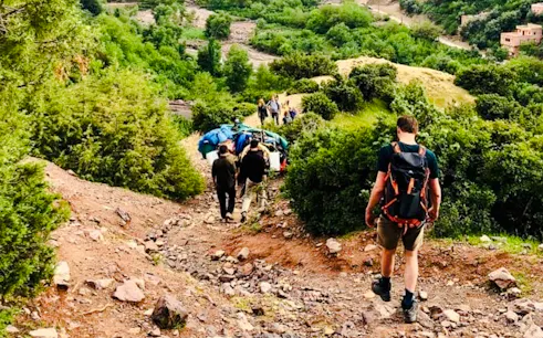 Hikers descending a rocky trail in the Al Haouz Mountains, Marrakesh, surrounded by lush greenery.