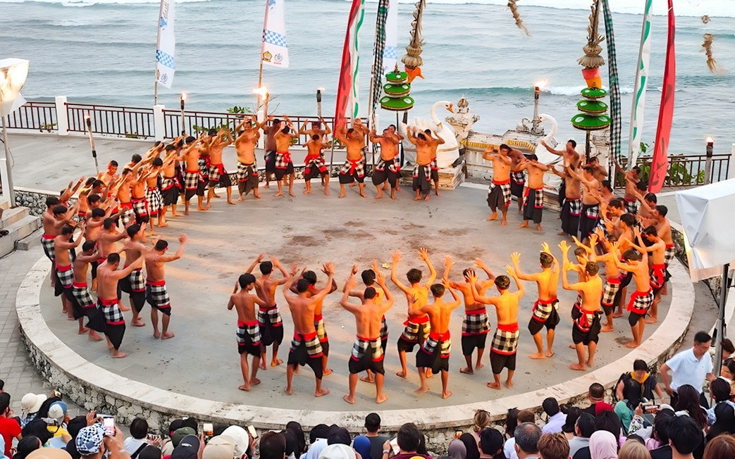 Kecak dancers performing at Melasti Beach amphitheater with ocean backdrop.