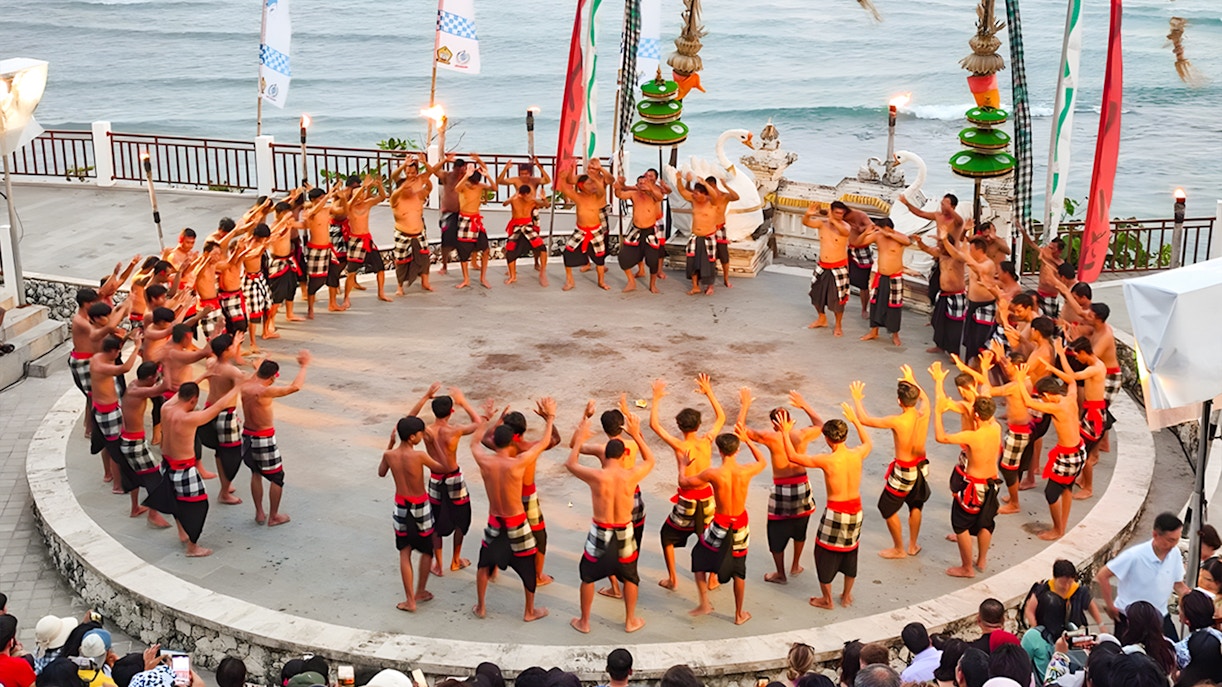 Kecak dancers performing at Melasti Beach amphitheater with ocean backdrop.