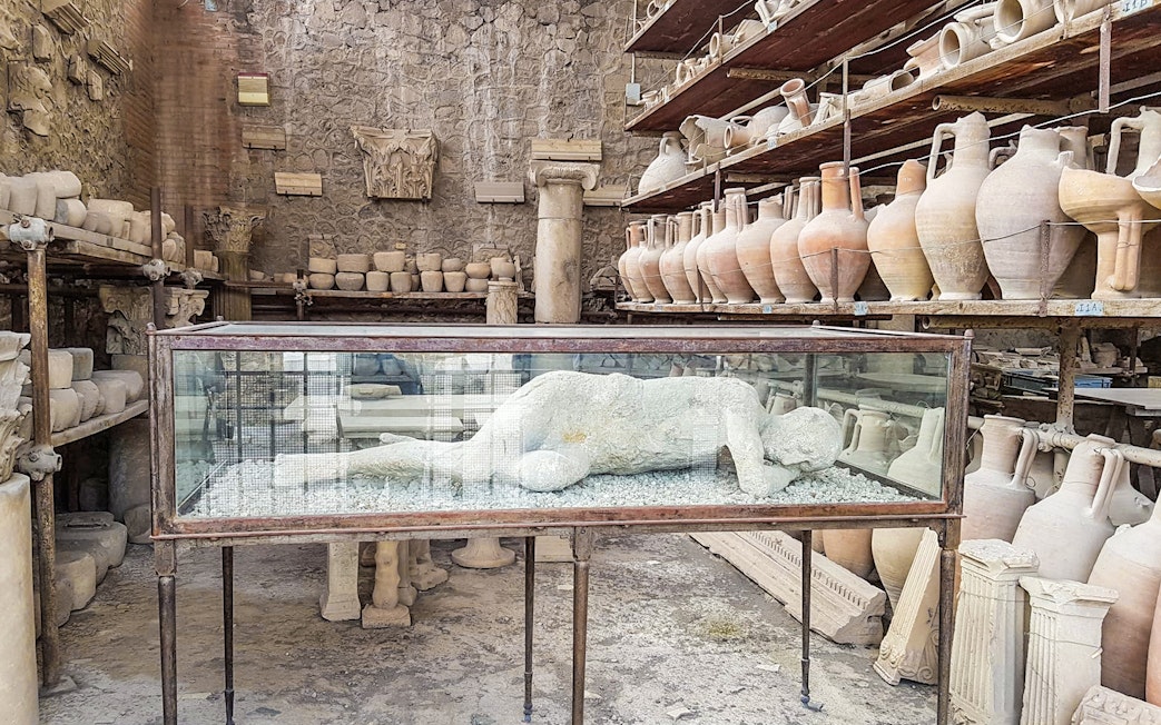 Plaster cast of a Pompeii inhabitant in a glass case surrounded by ancient pottery.