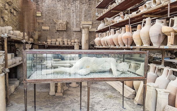 Plaster cast of a Pompeii inhabitant in a glass case surrounded by ancient pottery.