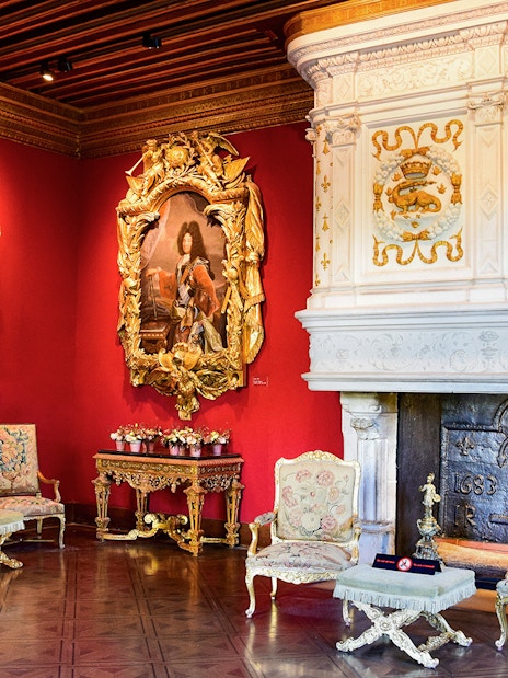 Ornate room with red walls, classic paintings, and a decorative fireplace in a historic European palace.