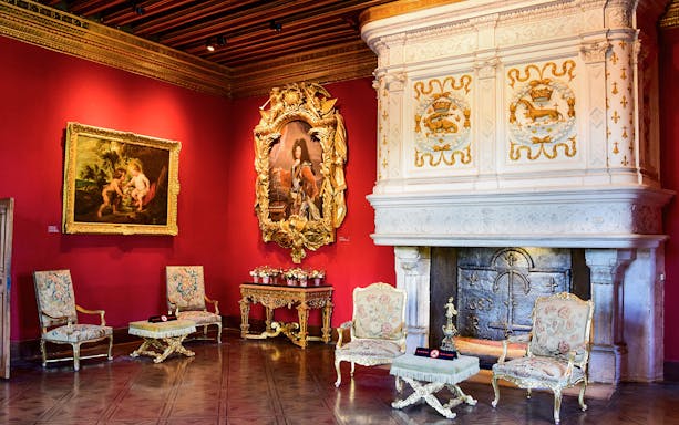 Ornate room with red walls, classic paintings, and a decorative fireplace in a historic European palace.