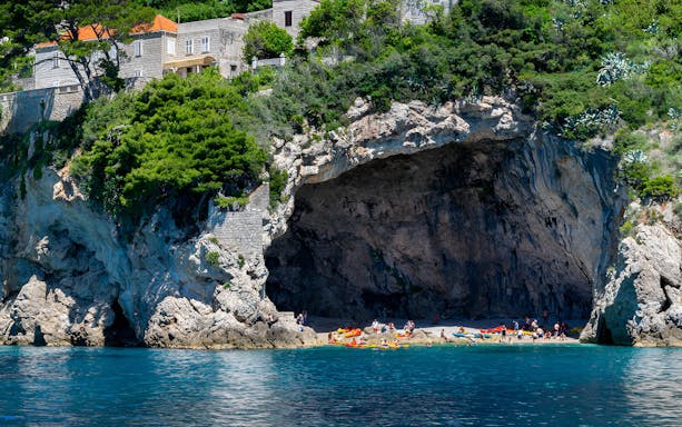 Kayakers and visitors at Betina Cave Beach, Dubrovnik, with rocky cliffs and lush greenery.
