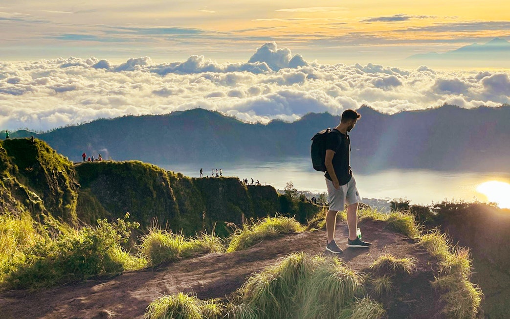 Man trekking Mount Batur at sunrise, overlooking clouds and distant peaks.