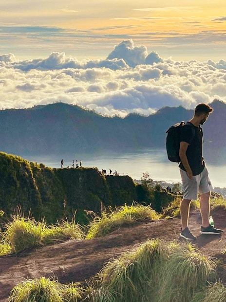 Man trekking Mount Batur at sunrise, overlooking clouds and distant peaks.