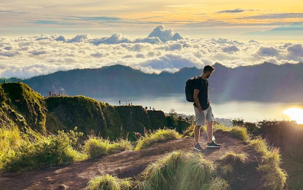 Man trekking Mount Batur at sunrise, overlooking clouds and distant peaks.