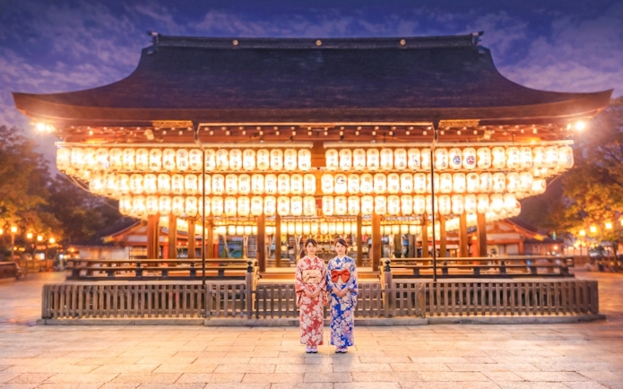 Yasaka Shrine illuminated at night in Kyoto, Japan, with two people in traditional attire.