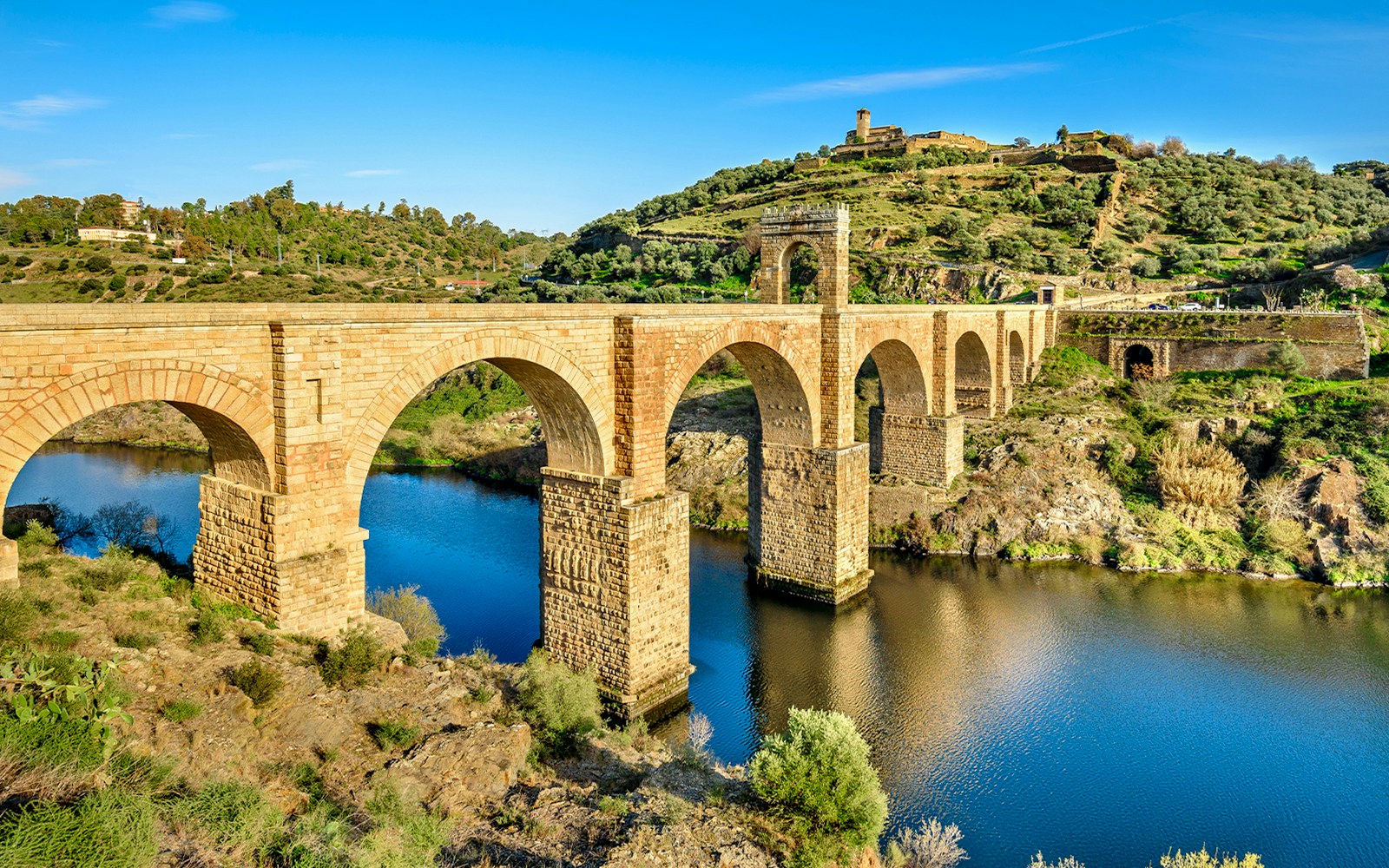Alcántara Roman Bridge spanning the Tagus River in Toledo, surrounded by lush landscape.
