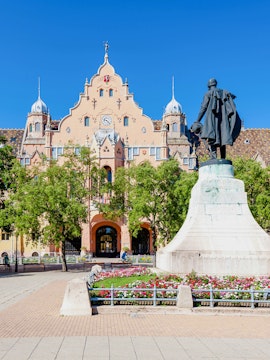 Town hall in Kecskemét, Hungary with a statue and flower garden in the foreground.