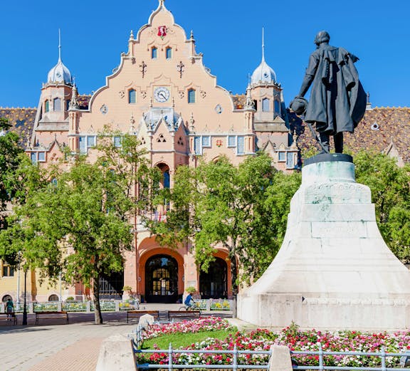 Town hall in Kecskemét, Hungary with a statue and flower garden in the foreground.