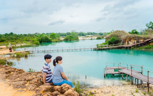 Couple sitting by Blue Lake, Bintan Mini Desert, with wooden pier and lush greenery.