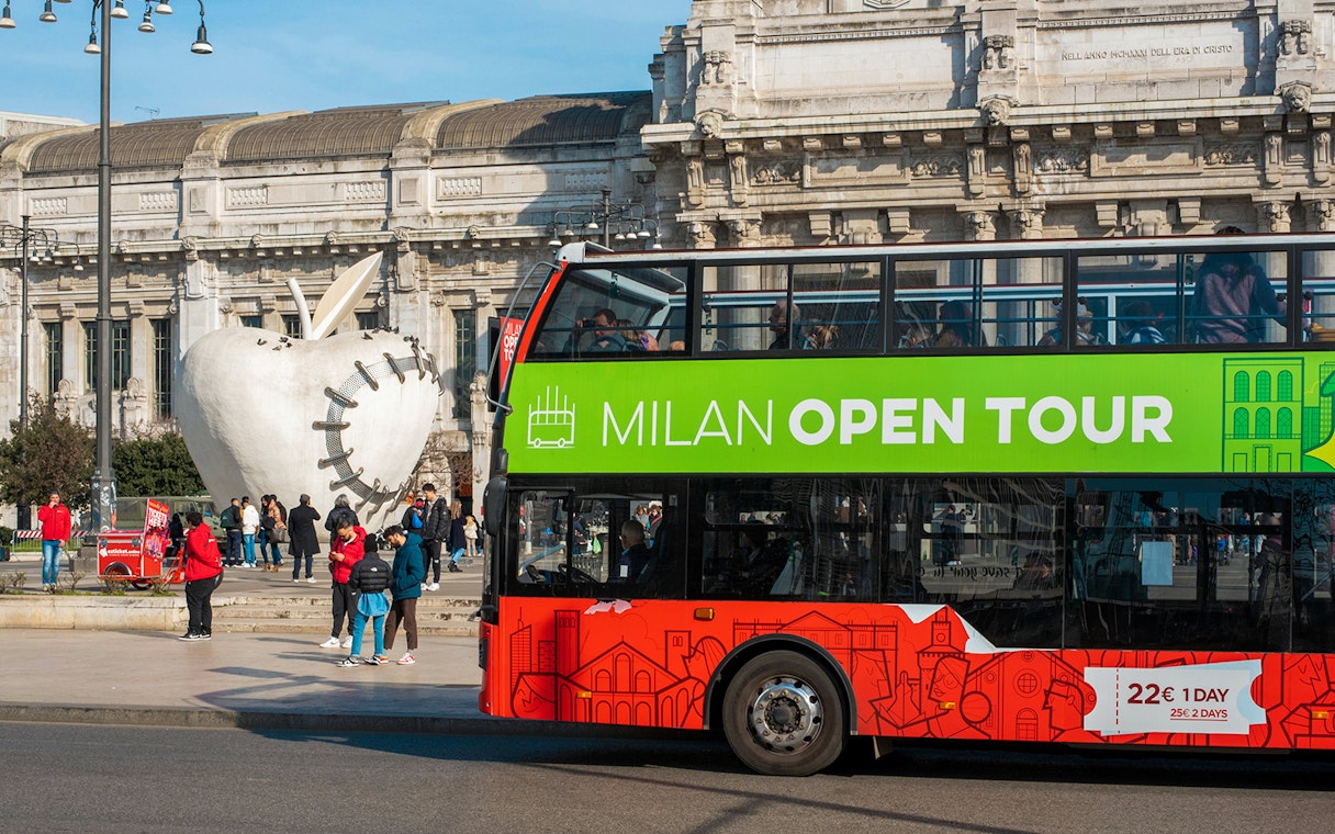 Milan hop-on hop-off tour bus near Central Station and apple sculpture.