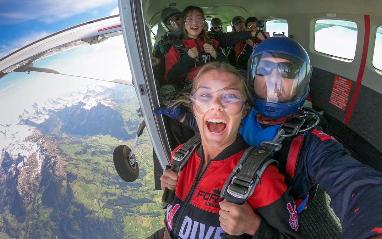 Tourists preparing to skydive from a plane over scenic mountains.