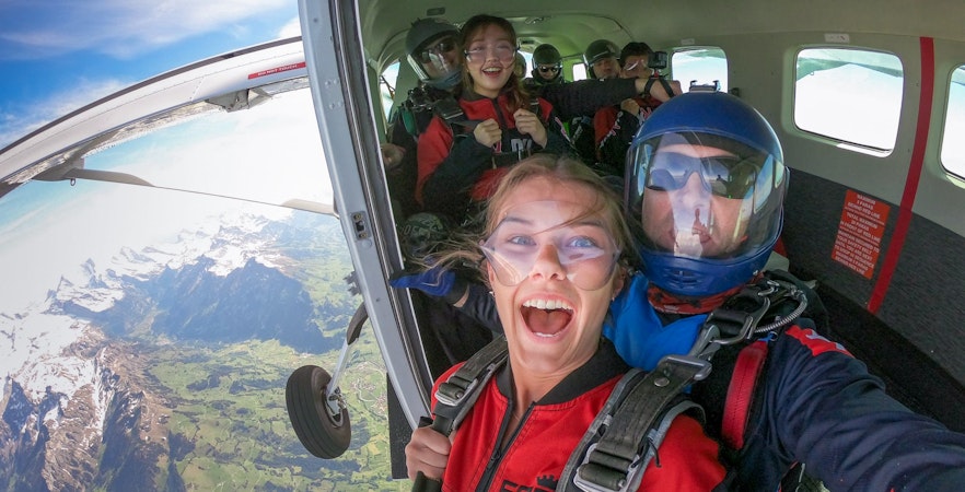 Tourists preparing to skydive from a plane over scenic mountains.