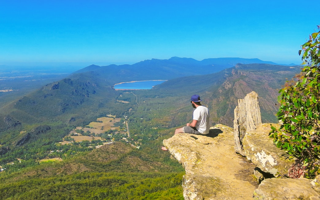 Person sitting on a rock ledge overlooking Grampians National Park landscape, Melbourne day tour.