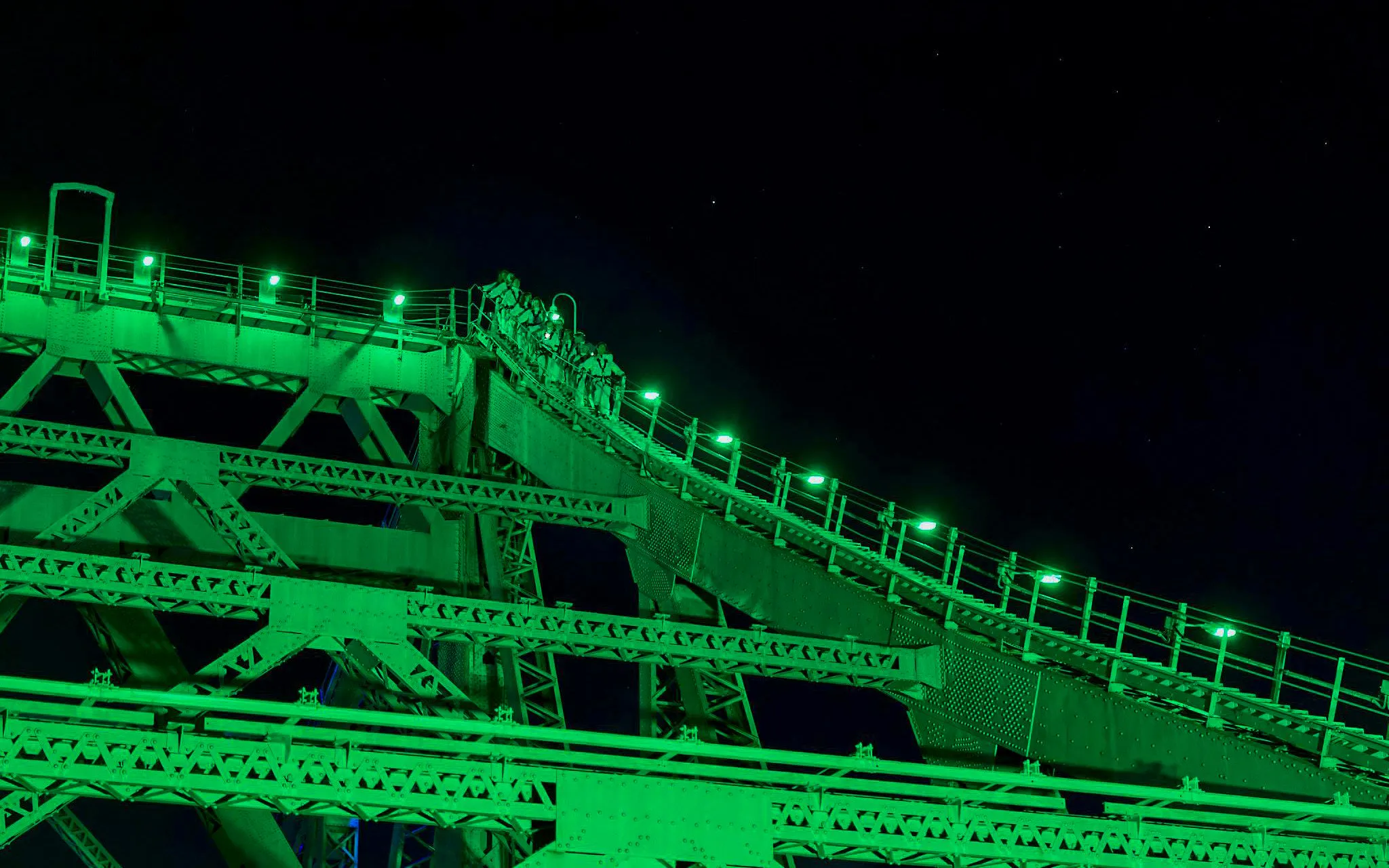 Climbers ascending Story Bridge at night, illuminated by green lights in Brisbane.