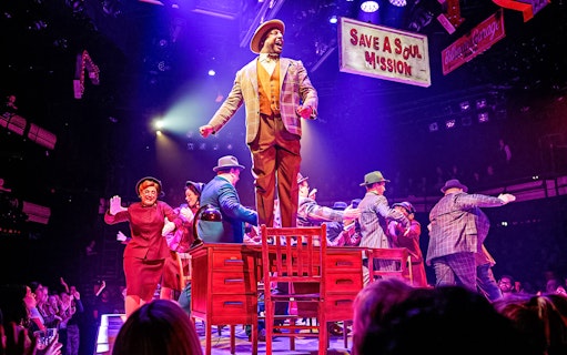 Stage performance of "Guys & Dolls" with actors dancing on a table under colorful lights.
