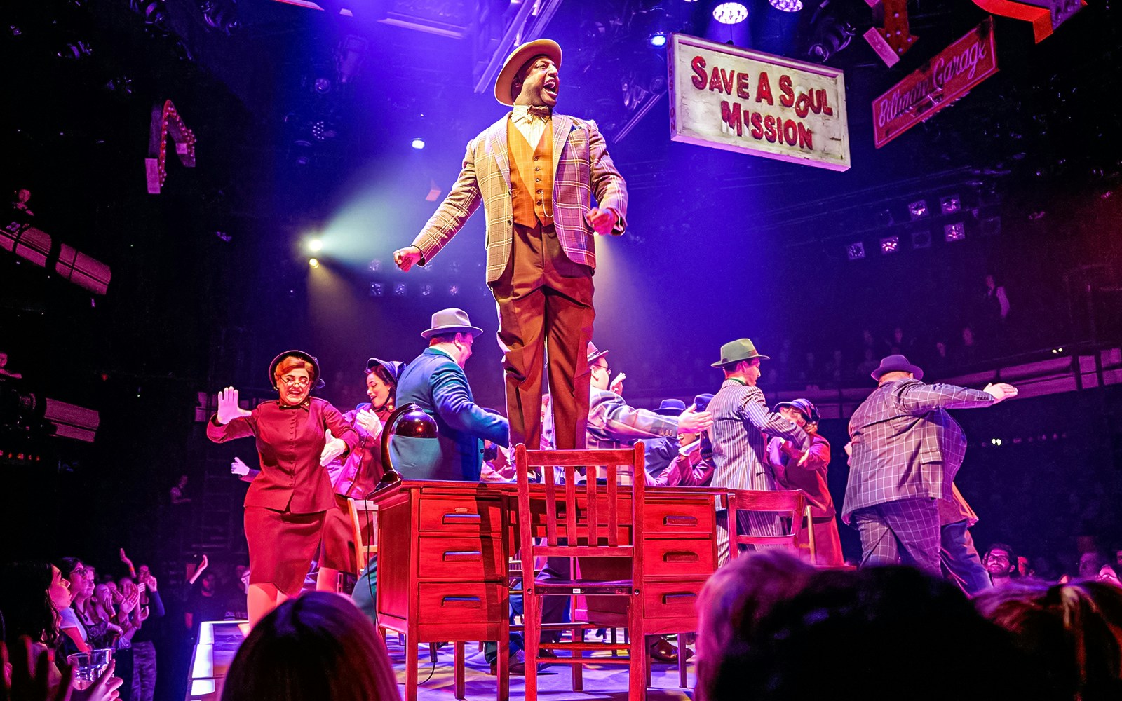 Stage performance of "Guys & Dolls" with actors dancing on a table under colorful lights.