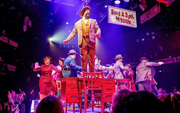 Stage performance of "Guys & Dolls" with actors dancing on a table under colorful lights.