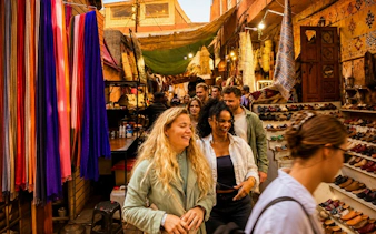 Tourists exploring a vibrant souk in Marrakech's Medina with colorful textiles and shoes displayed.