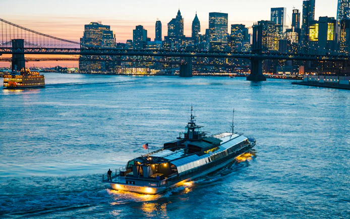 Dinner cruise boat on New York's East River with city skyline and bridge at sunset.