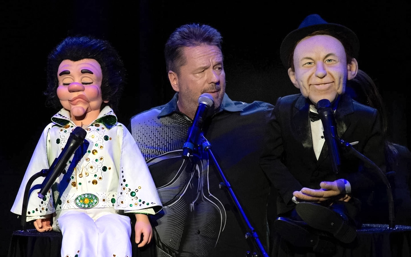 Ventriloquist performing with Elvis and Frank Sinatra puppets on stage.
