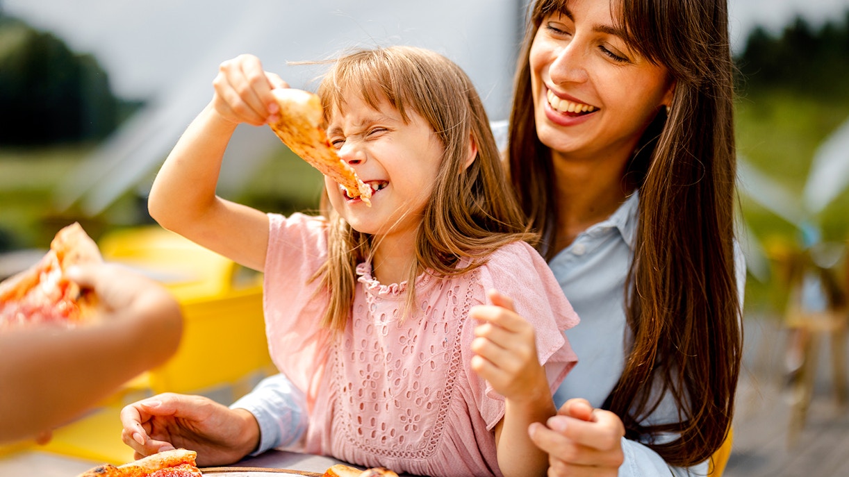 Family enjoying pizza outdoors, child smiling with a slice in hand.