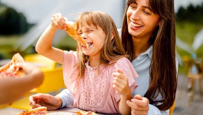 Family enjoying pizza outdoors, child smiling with a slice in hand.