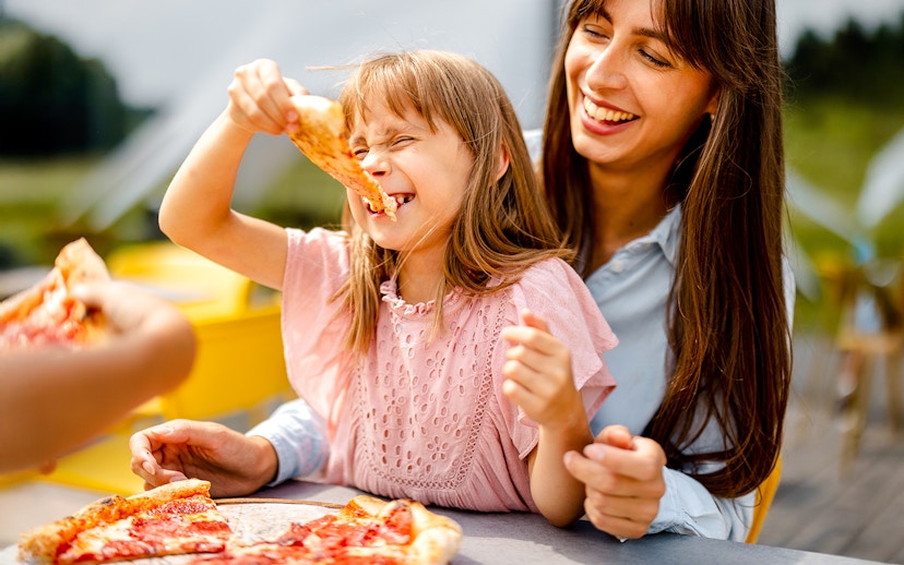 Family enjoying pizza outdoors, child smiling with a slice in hand.