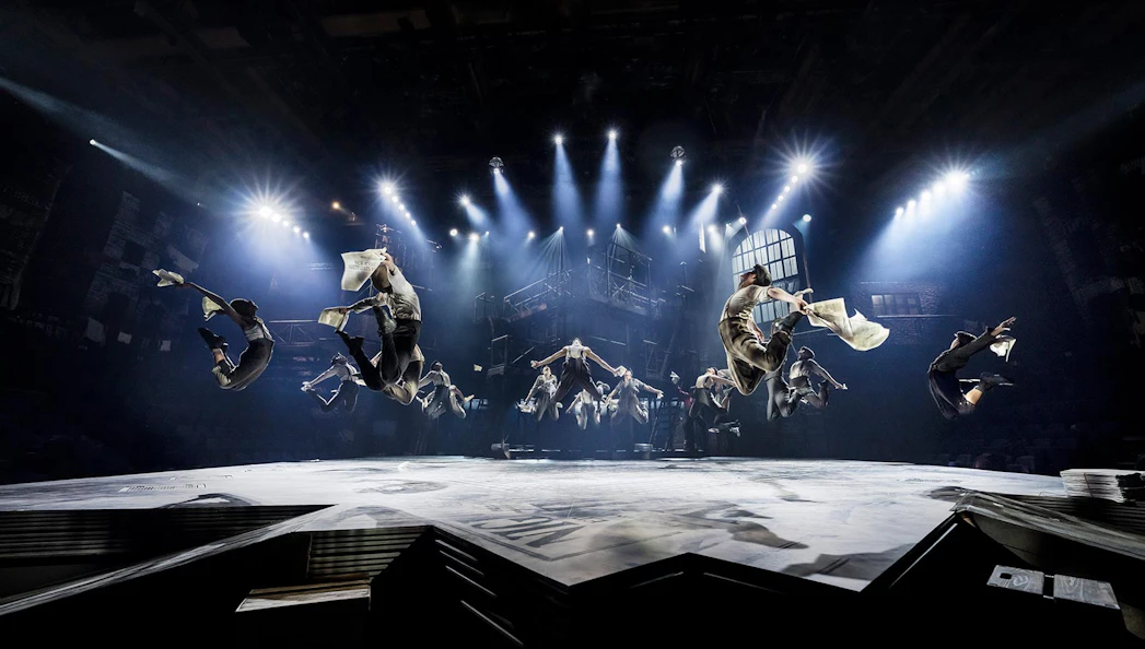 Performers leaping with newspapers in a dramatic scene from Newsies musical.