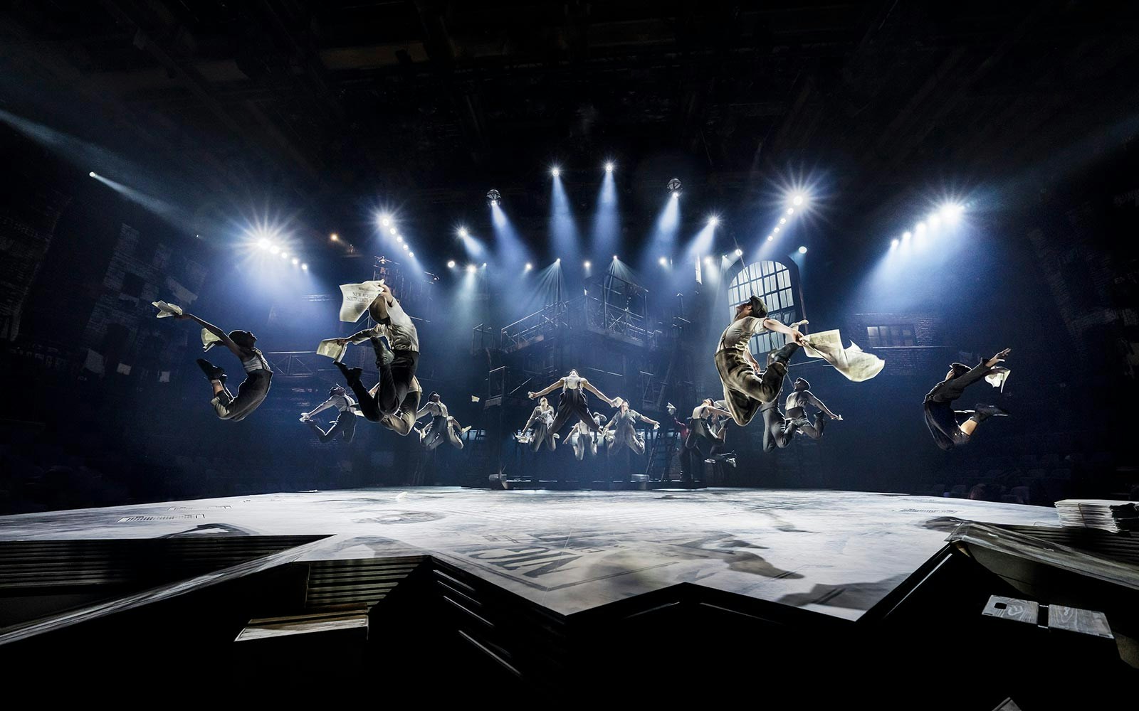 Performers leaping with newspapers in a dramatic scene from Newsies musical.